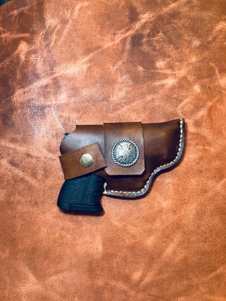 Op de afbeelding: Een bruine leren holster met een zilveren concho en een zwart pistool erin.