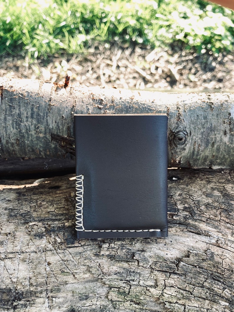 Puede incluir: Una cartera de cuero marr&oacute;n oscuro con costuras blancas. La cartera est&aacute; cerrada y se encuentra sobre una pieza de madera desgastada.