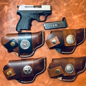 Op de afbeelding: Vier bruine leren holster met zilveren accenten en decoratieve ontwerpen. De holster zijn ontworpen om een klein, zilveren pistool te dragen.