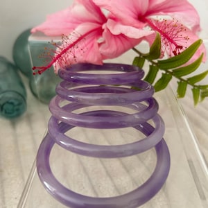 May include: Stack of six translucent lavender bangles. A pink hibiscus flower with a red stamen and green leaves are in the background. The bangles are stacked on a clear acrylic surface.