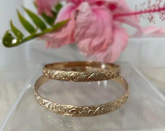 Puede incluir: Dos brazaletes dorados con intrincados diseños florales y de espirales. Los brazaletes están apilados, reflejando la luz. Una flor de hibisco rosa y hojas verdes en el fondo. Los brazaletes miden aproximadamente 6,5 cm de diámetro.