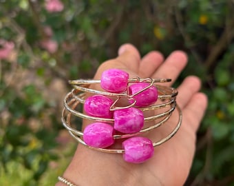 Puede incluir: Un conjunto de brazaletes de oro con cuentas de ágata rosa y blanca. Los brazaletes están apilados uno encima del otro y se llevan en la muñeca.