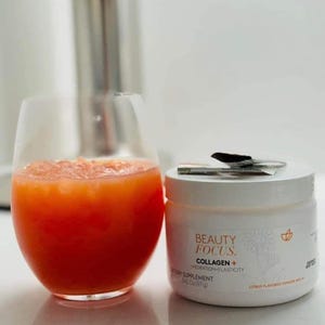 Puede incluir: Un tarro blanco de suplemento diet&eacute;tico en polvo de col&aacute;geno Beauty Focus+ con sabor a c&iacute;tricos. El tarro est&aacute; sobre una superficie blanca junto a un vaso de zumo de naranja.