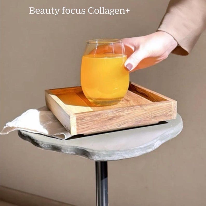 Puede incluir: Un vaso de zumo de naranja en una bandeja de madera, sobre una mesa de piedra gris. El texto "Beauty focus Collagen+" es visible sobre el vaso.