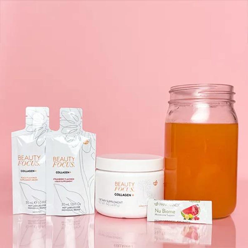 Puede incluir: Un conjunto de suplementos de belleza que incluye un tarro de polvo de col&aacute;geno, dos paquetes individuales de col&aacute;geno y un paquete de apoyo a la hidrataci&oacute;n Nu Biome. El polvo de col&aacute;geno es blanco con las palabras "Beauty Focus Collagen+" impresas en el tarro. Los paquetes son blancos con las palabras "Beauty Focus Collagen+" impresas en ellos. El paquete Nu Biome es blanco con las palabras "Nu Biome Moisture Support" impresas en &eacute;l. El fondo es rosa claro.
