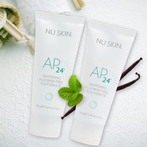 May include: Two white tubes of Nu Skin AP24 Whitening Fluoride-Free Toothpaste. The tubes are labeled with the product name and have a green and white design. The left tube has a sprig of mint leaves and a vanilla bean next to it. The right tube has a vanilla bean next to it.