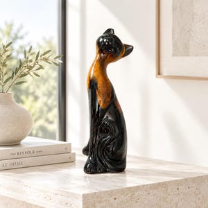 May include: A tall, stylised ceramic cat figurine featuring a glossy finish with a gradient colour scheme transitioning from deep black at the base and head to a warm, mottled amber-orange tone across the chest and neck, posed in a seated position.