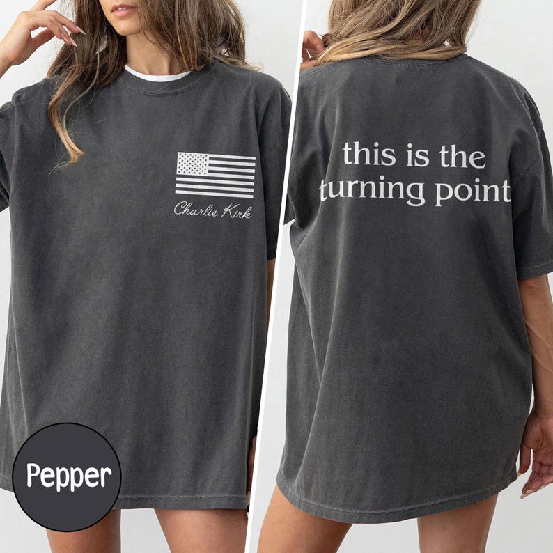 May include: Pepper-colored t-shirt with a white American flag graphic and the name "Charlie Kirk" printed on the front. The back of the shirt features the text "this is the turning point" in white. The shirt has a relaxed fit.