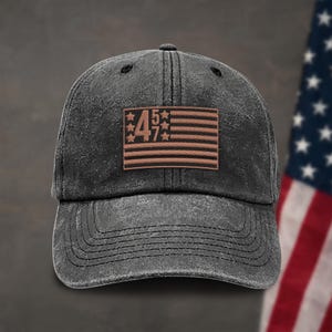 K&ouml;nnte beinhalten: Eine schwarze, abgenutzte Baseballkappe mit einem braunen Patch, der eine stilisierte amerikanische Flagge mit den Zahlen 457 in den Sternen zeigt.