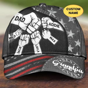 May include: A black baseball cap with a design featuring fists and the words "DAD," "ADDIE," "MAX," "JACK," "LAYLA," and "MILO." The cap also has a "CUSTOM NAME" label and the text "Best Grandpa Ever."