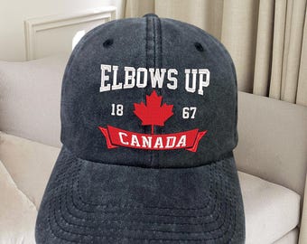 Pode incluir: Um boné de beisebol preto com um logotipo bordado branco que diz "Elbows Up 18 67 Canada" com uma folha de bordo vermelha.