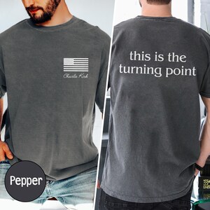 May include: A pepper-colored t-shirt with a white American flag graphic and the text "Charlie Kirk" on the front. The back of the shirt features the phrase "this is the turning point" in white.