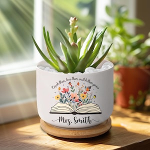 May include: A white ceramic planter with a succulent plant, featuring the text "Teach them, love them, watch them grow" and "Mrs. Smith". The planter has a floral design and sits on a wooden coaster.