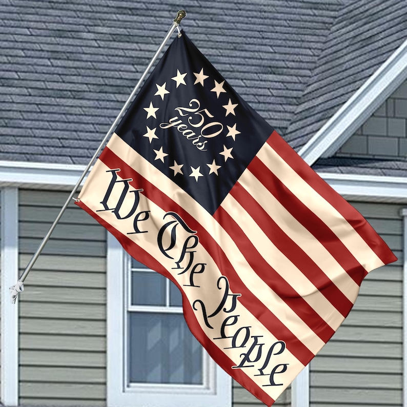 May include: A patriotic flag with a design reminiscent of the American flag, featuring red and white stripes and a blue field with stars. The flag displays the text "We The People" and "250 years". It is mounted on a silver pole and displayed outside a house.