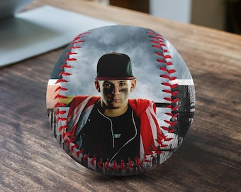 May include: A baseball with a red and white stitching pattern. The baseball features a photo of a person wearing a black baseball cap and a red and white jacket. The background of the photo is a blurry image of a baseball field.