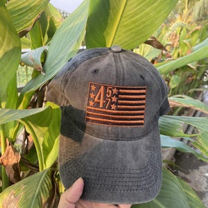 K&ouml;nnte beinhalten: Eine schwarze Baseballkappe aus Denim mit einer gestickten amerikanischen Flagge und den Zahlen "45" und "7" in Orange.