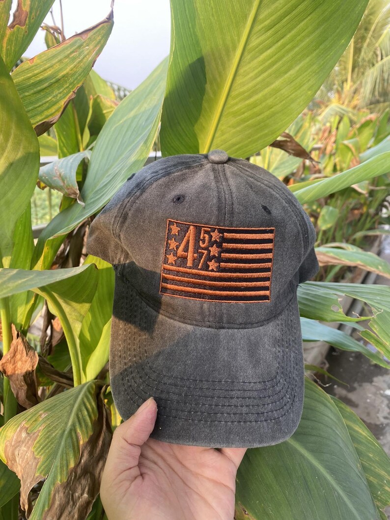 K&ouml;nnte beinhalten: Eine schwarze Baseballkappe aus Denim mit einer gestickten amerikanischen Flagge und den Zahlen "45" und "7" in Orange.