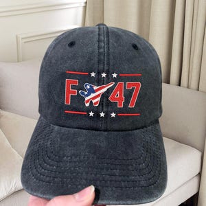May include: A black denim baseball cap with red and white embroidery. The embroidery features a stylized American flag with a jet fighter plane and the text "F-47".