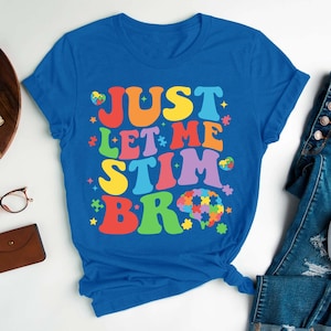 May include: A royal blue t-shirt with the text "JUST LET ME STIM BR" in rainbow colors. The text is in a retro font style. A puzzle piece brain is included in the design.