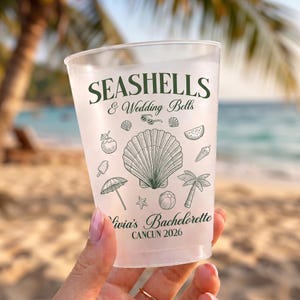 May include: A frosted plastic cup featuring dark green text that reads SEASHELLS & Wedding Bells, Olivia's Bachelorette, and CANCUN 2026. The design includes line art illustrations of a large seashell, palm tree, umbrella, and beach items.