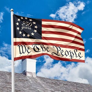 May include: A flag with a design featuring red and white stripes and a blue canton with stars. The flag reads "250 years" and "We The People" in a decorative font. The flag is waving in the wind against a blue sky.