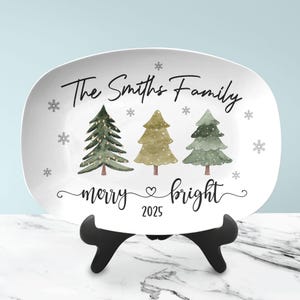 May include: White rectangular serving platter with a Christmas tree design. The platter features three watercolor Christmas trees, snowflakes, and the text "The Smiths Family", "merry & bright", and "2025". The platter is displayed on a black stand.
