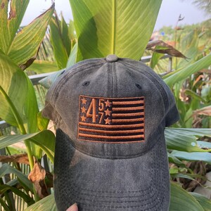 K&ouml;nnte beinhalten: Eine schwarze und braune Baseballkappe mit einem gestickten amerikanischen Flaggenmuster mit den Zahlen "457" in der Mitte.