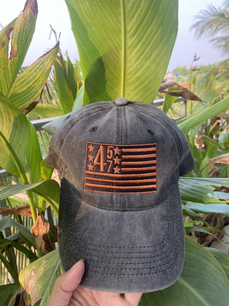 K&ouml;nnte beinhalten: Eine schwarze und braune Baseballkappe mit einem gestickten amerikanischen Flaggenmuster mit den Zahlen "457" in der Mitte.