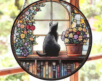 May include: A stained glass window hanging with a black cat sitting on a windowsill looking out a window. The windowsill has a bookshelf with books below it. There are flowers in a pot on the right side of the window.