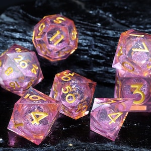 Könnte beinhalten: Ein Set aus sieben durchscheinenden, rosa Würfeln mit goldenen Zahlen und Glitzer. Die Würfel haben verschiedene polyedrische Formen, darunter ein d20, d12, d10, d8, d6 und d4, geeignet für Tabletop-Rollenspiele.