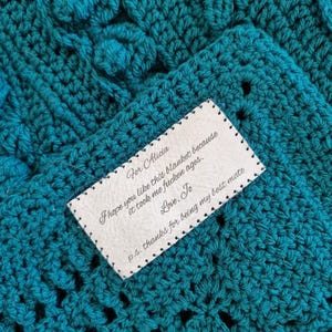 May include: A teal crocheted blanket with a textured floral design. A white tag with black text is sewn onto the blanket. The text reads: "For Alicia, I hope you like this blanket because it took me fucken ages. Love, To, p.s. thanks for being my best mate."