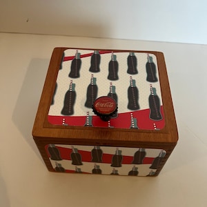 May include: A wooden box with a Coca-Cola themed design. The lid features a pattern of Coca-Cola bottles with red and white stripes. The box has a Coca-Cola bottle cap as a knob.