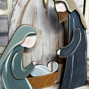 May include: A wooden nativity scene featuring Mary, Joseph, and baby Jesus. The figures are painted in muted tones of blue, green, and brown, with a star above. The scene is set against a weathered wooden backdrop.