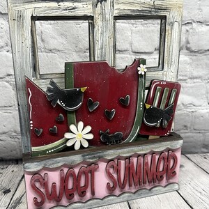 May include: A decorative wooden sign featuring a distressed white window frame, a red watermelon slice, a popsicle, black birds, white daisies, and small hearts. The base is a pink rectangular box with the red text SWEET SUMMER.