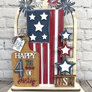 May include: A patriotic wooden decoration featuring the American flag with red and white stripes and blue field with white stars. The words "Happy 4th of July" are displayed on a wooden plaque, along with the word "USA". Decorative stars and fireworks complete the design.