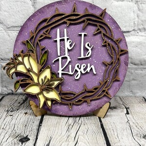 May include: A round, purple Easter decoration with the text "He Is Risen" in white. The design includes a brown crown of thorns and a cluster of yellow and green lilies. The decoration is on a wooden stand.