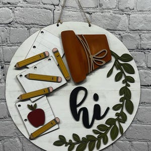 May include: A round, white wooden sign with school-themed decorations. It features pencils, notebooks, a red apple, and a brown book. The word "hi" is written in black script, and green leaves surround the bottom.