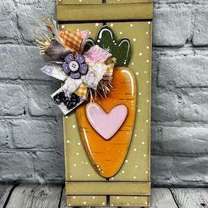 May include: Wooden carrot-shaped wall decor with a pink heart, green leaves, and a decorative fabric bow. The carrot is orange with white accents, set against a beige background with white polka dots. A charming Easter or spring decoration.