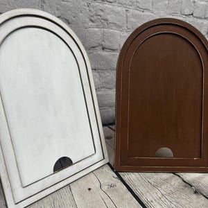 May include: Two wooden arched frames, one painted white and the other stained brown. Each frame has a recessed panel and a semi-circular cutout at the bottom. The frames are set against a brick wall and a wooden surface.