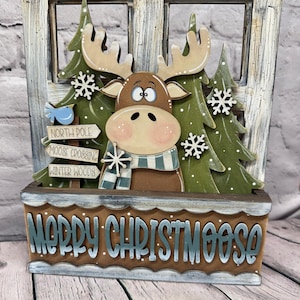 May include: A wooden decorative sign featuring a brown moose wearing a blue and white striped scarf, positioned in front of green pine trees and a window frame. The base displays the text MERRY CHRISTMOOSE, with signs reading NORTH POLE, MOOSE CROSSING, and WINTER WOODS.