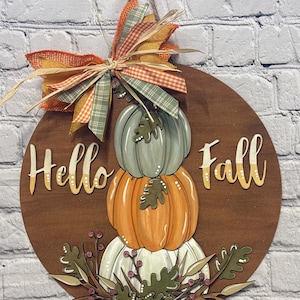 May include: A round wooden fall-themed sign with the words "Hello Fall" in a script font. The sign features a stack of three pumpkins in shades of green, orange, and white, with a decorative bow at the top. The bow has orange, green, and tan ribbons.