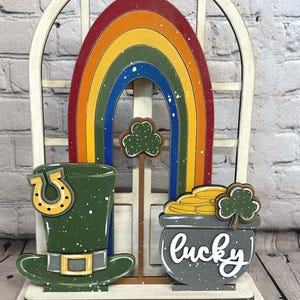 May include: A decorative wooden display featuring a white arched window frame backdrop. In front, a colorful rainbow, a green leprechaun hat with a yellow horseshoe, a grey pot filled with gold coins, and two green shamrocks are arranged.