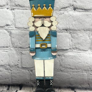 May include: A wooden nutcracker figurine in shades of blue, gold, and white. The figurine has a crown, curly hair, a mustache, and a beard. It is wearing a blue uniform with a gold belt and black boots. The nutcracker stands on a black base.