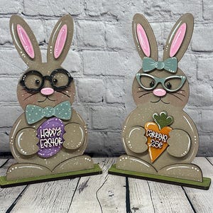 May include: Two wooden Easter bunny decorations. One bunny wears glasses and a bow tie and holds a purple egg with "Happy Easter" on it. The other bunny wears glasses and a bow and holds a carrot with "Carrots 25¢" on it. Both bunnies have pink ears.