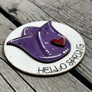 May include: A round wooden decoration with a purple flower design and a small red heart. The words "HELLO SPRING" are written in black script around the edge of the white circle. The background is a wooden surface.