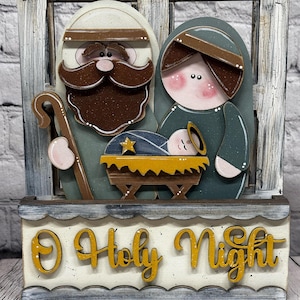 May include: Wooden Christmas nativity scene featuring Joseph, Mary, and baby Jesus in a manger. The figures are set against a window frame with the words "O Holy Night" in yellow script. The overall design has a rustic, hand-painted aesthetic.