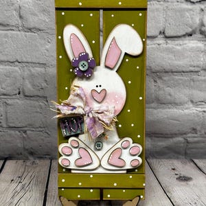 May include: A vertical olive green wooden shutter sign with white polka dots featuring a white bunny cutout. The bunny has pink ears, a heart-shaped nose, and pink paw pads. It is decorated with a purple flower, a fabric bow, and a sign reading HOP.