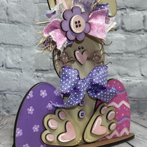 May include: A decorative wooden bunny cutout featuring a tan body, a purple polka dot bow, and a large purple flower accent. The bunny is flanked by two painted wooden eggs, one purple with small flowers and one pink with wavy stripes.