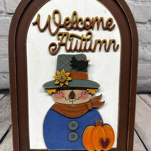 May include: Wooden autumn-themed decoration featuring a scarecrow design. The sign reads "Welcome Autumn" in cursive. The scarecrow has a blue shirt, a hat, and an orange pumpkin. The frame is brown.