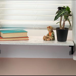 May include: A white shelf with black brackets is mounted above a window. The shelf is decorated with a small figurine of a girl reading a book, a potted plant, and three books.